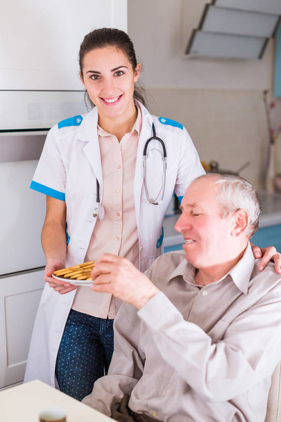 De gelukkige jonge dokter permanent in de buurt van oude, zieke man en cookies hem in de keuken - Foto, afbeelding