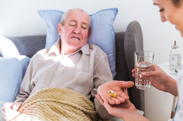 The old sick man lying on a bed and a doctor sitting near him and giving pills and water him - Photo, Image