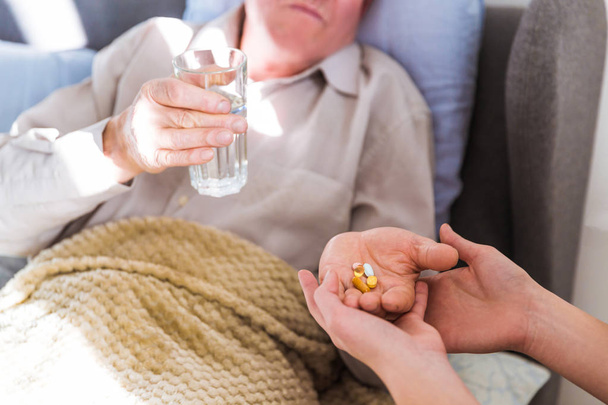 Close-up the old sick man lying on a bed at home and holding pills and water in hands to drink medicines - Foto, imagen