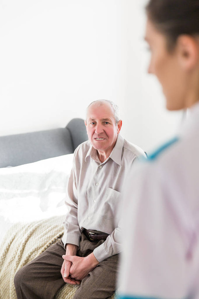 The old smiling man sitting on a bed in the room at home and young nurse entering in a room - Photo, image