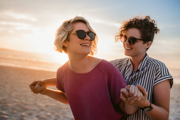 Carefree young lesbian couple wearing sunglasses playfully dancing hand in hand together on a sandy beach at sunset - Photo, image