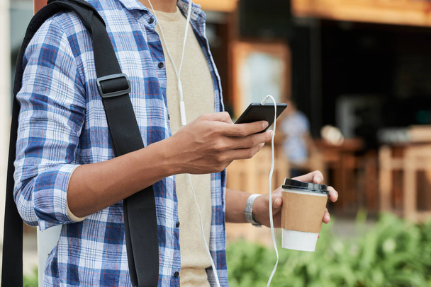 Unbekannter Mann mit T-Shirt und kariertem Hemd surft auf Smartphone im Internet und trinkt duftenden Kaffee im Freien, verschwommener Hintergrund - Foto, Bild