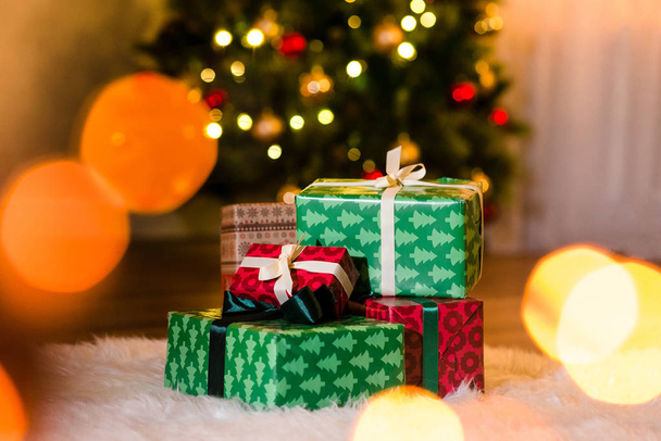 Несколько красивых подарков в новогодней оберточной бумаге на пушистом белом ковре, красивая елка на заднем плане. Рождественский праздник, украшенный праздничными шарами, копирайт
 - Фото, изображение