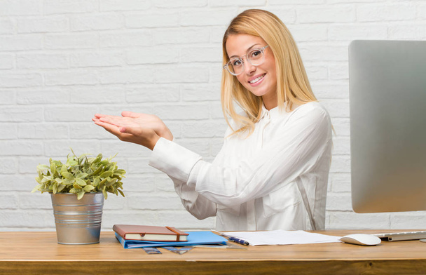 Portrait d'une jeune étudiante assise sur son bureau faisant des tâches tenant quelque chose avec les mains, montrant un produit, souriante et joyeuse, offrant un objet imaginaire
 - Photo, image