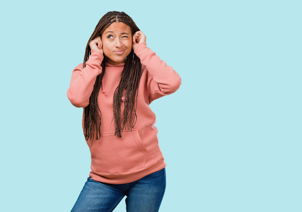 Retrato de una joven mujer negra con trenzas cubriendo los oídos con las manos, enojada y cansada de escuchar algún sonido
 - Foto, Imagen