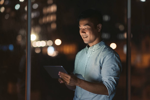 Smiling young Asian usinessman using a digital tablet while standing in an office late at night with city lights glowing in the background - 写真・画像
