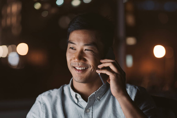 Smiling young Asian businessman talking on a cellphone while working in an office late at night with city lights glowing in the background - Фото, изображение
