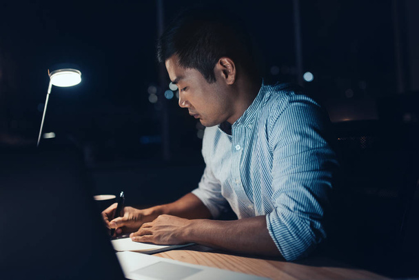 Young Asian businessman sitting at his desk, writing notes and working on a laptop in a dark office at night with city lights in the background - Photo, image