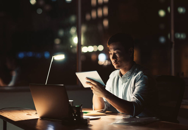 Young Asian businessman using a digital tablet while sitting at his desk in a dark office at night with city lights in the background - Fotografie, Obrázek