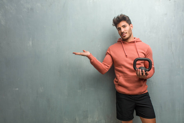 junger Fitnessmann vor einer Grunge-Wand, der etwas mit Händen hält, ein Produkt zeigt, lächelnd und fröhlich, ein imaginäres Objekt anbietet. mit einer eisernen Hantel. - Foto, Bild