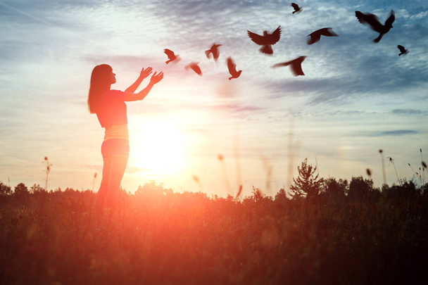 Una joven reza mientras disfruta de la naturaleza en medio de una hermosa puesta de sol. El concepto de esperanza, fe, religión. Una bandada de pájaros vuela, símbolo de esperanza y libertad
. - Foto, imagen