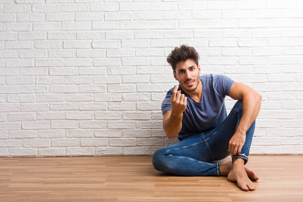 Young natural man sit on a wooden floor inviting to come, confident and smiling making a gesture with hand, being positive and friendly - Fotoğraf, Görsel