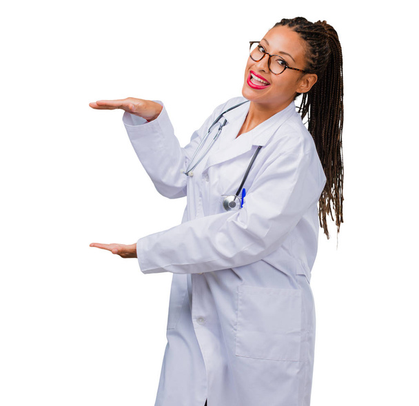 Portrait of a young black doctor woman holding something with hands, showing a product, smiling and cheerful, offering an imaginary object - Foto, Bild