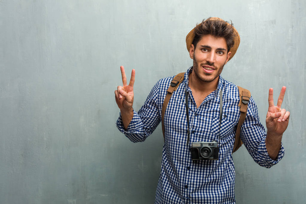 Jonge knappe reiziger man dragen van een hoed van stro, een rugzak en een fotocamera leuk en gelukkig, positieve en natuurlijke, doen een gebaar van overwinning, vrede-concept - Foto, afbeelding