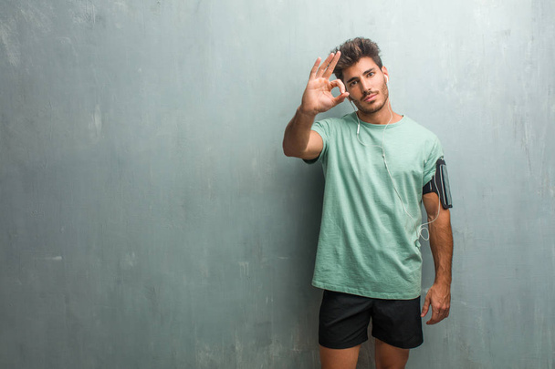 Young fitness man against a grunge wall cheerful and confident doing ok gesture, wearing an armband with phone. - Photo, Image