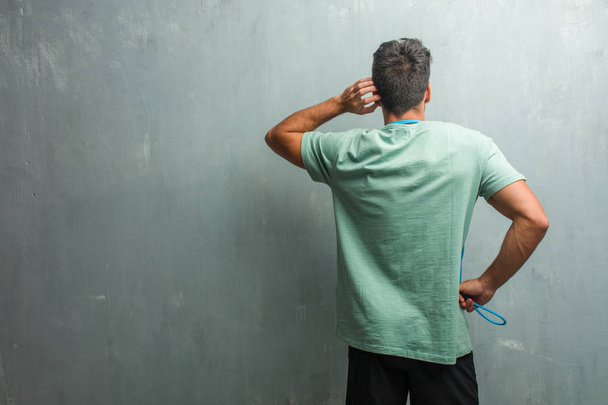 Young fitness man against a grunge wall showing back, posing and waiting, looking back. Holding a jump rope. - Photo, Image
