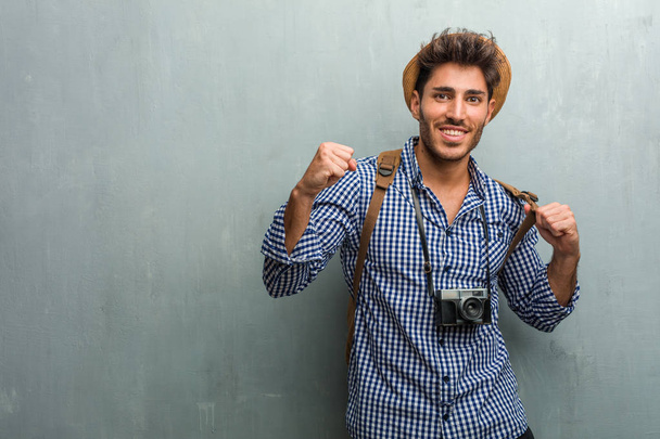 Jeune homme beau voyageur portant un chapeau de paille, un sac à dos et un appareil photo très heureux et excité, levant les bras, célébrant une victoire ou un succès, remportant la loterie
 - Photo, image
