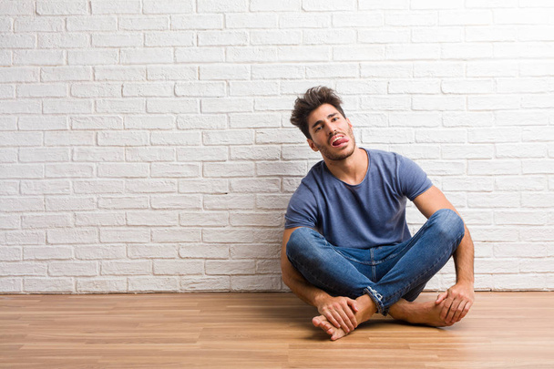 Young natural man sit on a wooden floor expression of confidence and emotion, fun and friendly, showing tongue as a sign of play or fun - 写真・画像