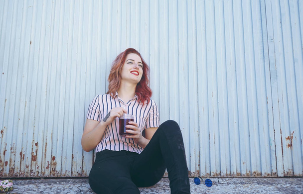Beautiful redhead woman drinking juice sitting against blue wall outdoor - Foto, Imagen
