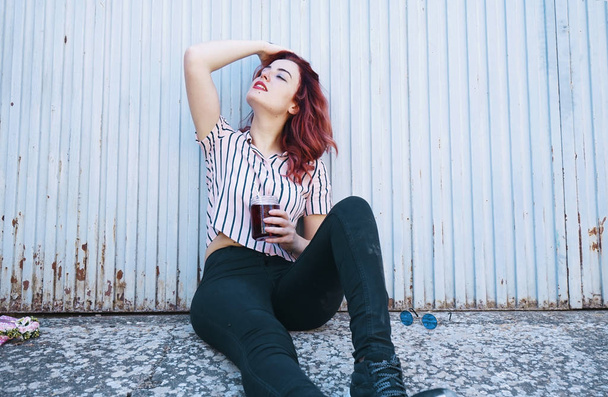 Piękne redhead kobieta picie soku, siedząc przed niebieską ścianą odkryty - Zdjęcie, obraz