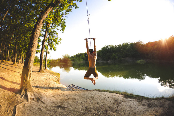 Salta del bungee. Un hombre está descansando en el lago. Un columpio de una cuerda y un palo. Recreación activa en la naturaleza. Diversión de verano. Lente de ojo de pez
. - Foto, imagen