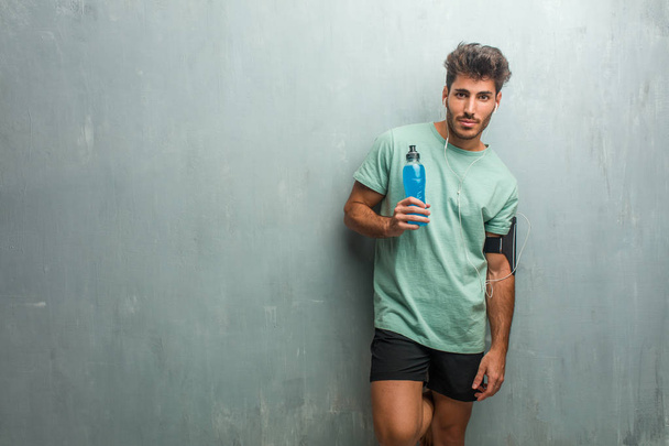 Junger Fitnessmann vor einer Grunge-Wand, fröhlich und mit einem breiten Lächeln, in der Hand einen blauen Energy-Drink. - Foto, Bild
