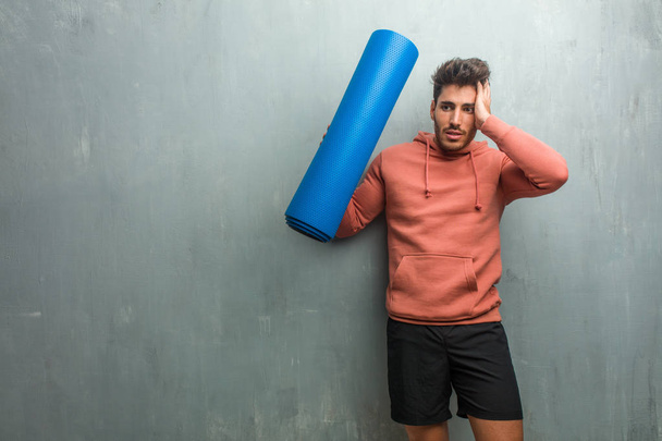 Człowiek młody fitness ścianę grunge, sfrustrowani i zdesperowani, trzymając niebieski Mata do uprawiania jogi. - Zdjęcie, obraz
