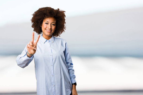 молода чорна жінка в синій сорочці робить жест перемоги на розмитому фоні
 - Фото, зображення