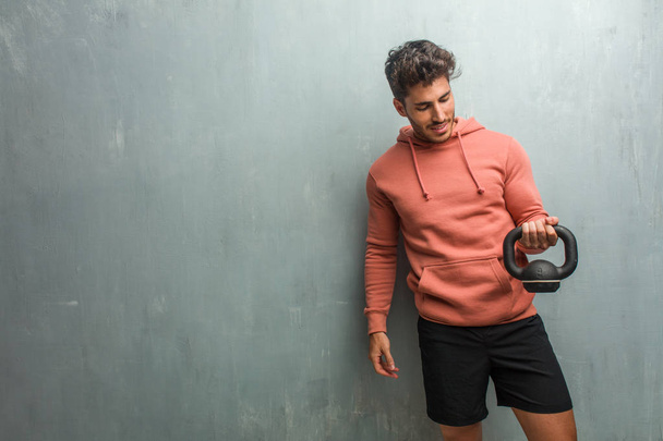 Young fitness man against a grunge wall laughing and having fun, being relaxed and cheerful, feels confident and successful. Holding an iron dumbbell. - Photo, Image