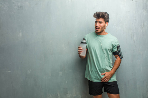 Young fitness man with sports bottle against a grunge wall very scared and afraid, wearing an armband with phone. - Photo, Image