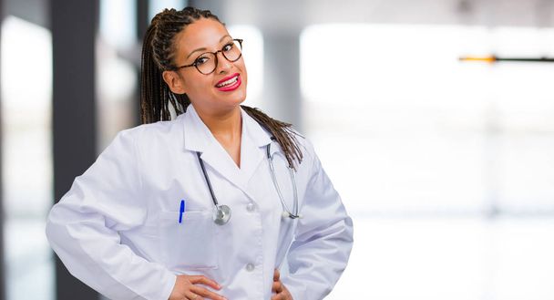 Porträt einer jungen schwarzen Ärztin mit den Händen auf den Hüften, stehend, entspannt und lächelnd, sehr positiv und fröhlich - Foto, Bild