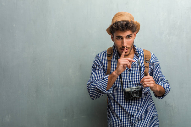Jonge knappe reiziger man met een hoed van stro, een rugzak en een fototoestel een geheimhouden of vragend om stilte, ernstige gezicht, gehoorzaamheid concept - Foto, afbeelding