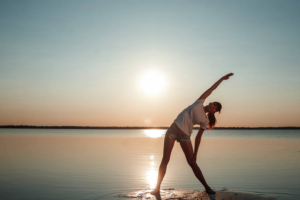 若い女の子のシルエット、湖と美しい夕日を背景に自然にヨガ。新鮮な空気、健康な生き方, - 写真・画像
