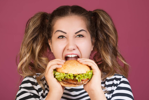 十代の少女は、分離のピンクの背景に幸せそうな顔と口を開けて空腹ビーフ ハンバーガー サンドイッチを保持しています。ファーストフードのコンセプト. - 写真・画像