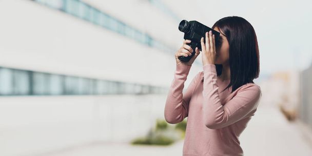 Young pretty woman excited and entertained, looking through a film camera, looking for an interesting shot, recording a movie, executive producer in the university campus - 写真・画像
