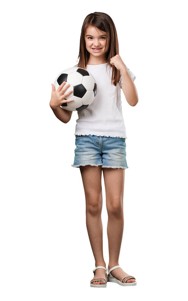Полное тело маленькая девочка улыбается и счастлива, держа в руках футбольный мяч, соревновательное отношение, с удовольствием играет в игру
 - Фото, изображение