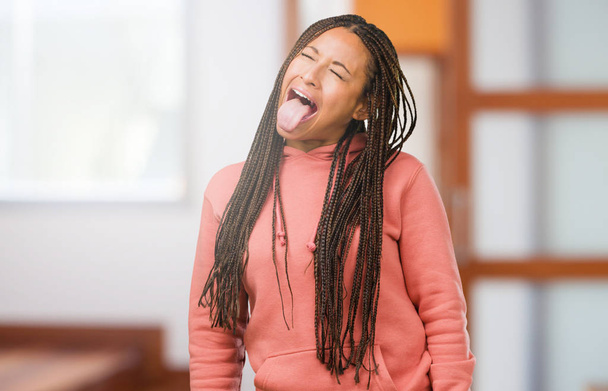 Πορτρέτο του μια νεαρή μαύρη γυναίκα φορώντας Τρέσες έκφραση της εμπιστοσύνης και συγκίνηση, διασκεδαστικό και φιλικό, δείχνοντας τη γλώσσα ως ένα σημάδι του παιχνιδιού και διασκέδασης - Φωτογραφία, εικόνα