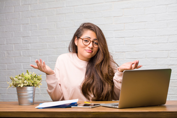 Portrait of young student latin woman sitting on desk doubting and shrugging shoulders - Foto, Bild