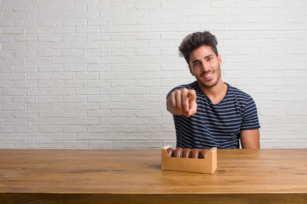 Young handsome and natural man sitting on a table cheerful and smiling pointing to the front. Eating chocolate doughnuts. - Photo, Image