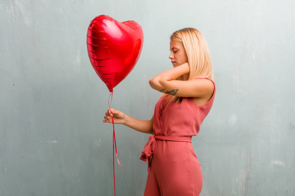 Portrait of young elegant blonde woman with back pain due to work stress, tired and astute. Holding a red heart balloon. - Fotografie, Obrázek
