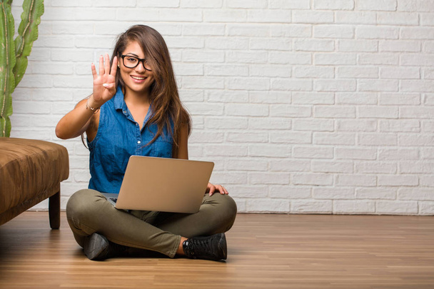 Portrait of young latin woman sitting on the floor showing the number four, symbol of counting, concept of mathematics, confident and cheerful. Holding a laptop. - Photo, image