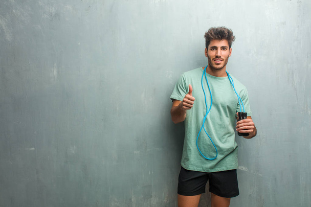 Young fitness man against a grunge wall cheerful and excited raising thumb up and holding a jump rope. - Photo, Image