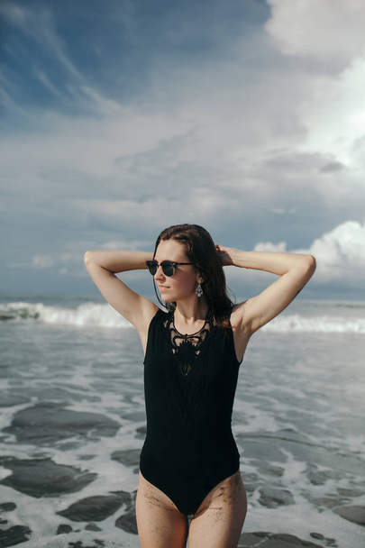 Feliz sonrisa morena Mujer con traje de baño negro en el hermoso cielo y el fondo del océano en la playa con arena negra. Bali, Canggu, Indonesia
 - Foto, imagen