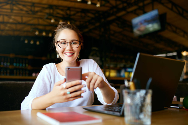Mixed race business woman typing a text on smartphone. Asian female holding a modern cellphone and writing message. Smiling young freelancer in glasses looking at camera with laptop on table. - Foto, immagini