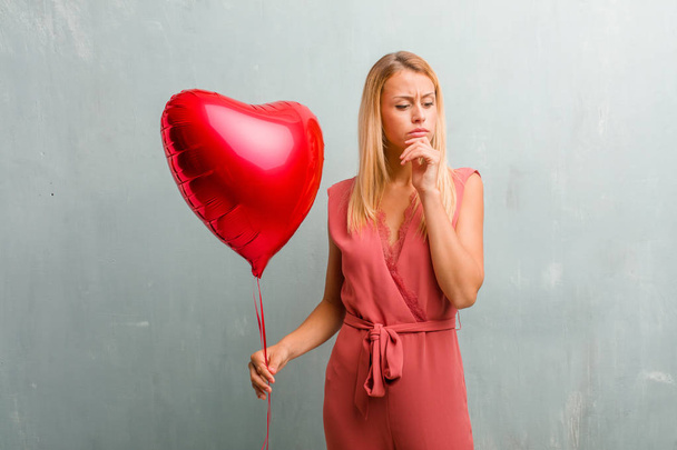 Portrait of young elegant blonde woman doubting and confused, thinking of an idea or worried about something. Holding a red heart balloon. - Photo, Image
