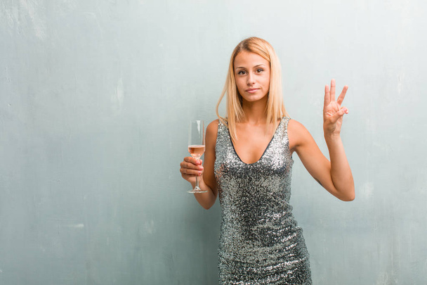 Porträt einer jungen eleganten blonden Frau, die Nummer drei zeigt, Symbol des Zählens, Konzept der Mathematik, selbstbewusst und fröhlich. in der Hand ein Champagnerglas. - Foto, Bild