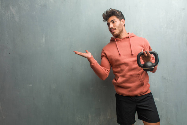 junger Fitnessmann vor einer Grunge-Wand, der etwas mit Händen hält, ein Produkt zeigt, lächelnd und fröhlich, ein imaginäres Objekt anbietet. mit einer eisernen Hantel. - Foto, Bild