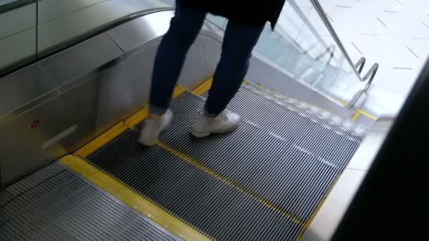 Une fille en chaussures légères et vêtements noirs vient sur les marches de l'escalator et descend. Les femmes nettoyantes en bas essuient le sol dans le centre commercial. Véritable prise de vue sans trépied latéral
. - Séquence, vidéo