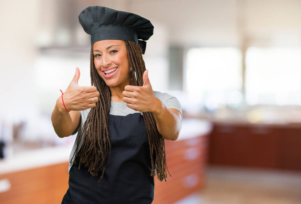 Nuoren mustan leipurin naisen muotokuva iloinen ja innoissaan, hymyillen ja nostaen peukalonsa ylös, menestyksen ja hyväksynnän käsite, ok ele
 - Valokuva, kuva