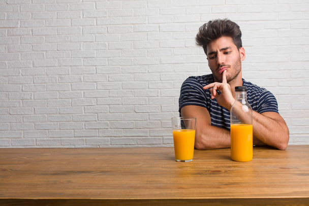 Young handsome and natural man sitting on a table doubting and confused, thinking of an idea or worried about something. Having a breakfast, includes orange juice and a cereals bowl. - Photo, Image
